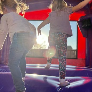 Operation Jump - Two children are playing inside an inflatable bounce house, jumping and reaching for the top. Sunlight streams through the window, creating a bright atmosphere. - Party Supplies Near Me - Leander, Texas