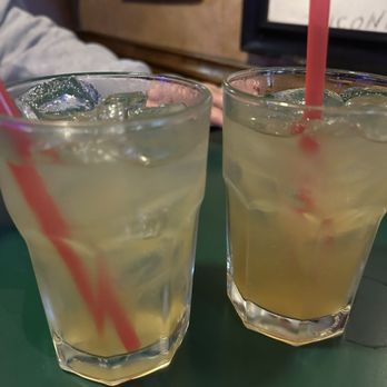 Harmon's Pub - Vegas bombs - Two glasses filled with yellowish drinks containing ice cubes and pink straws are positioned on a green table. The background shows part of a person’s arm and a wall with framed items, indicating a casual dining or bar environment. - Sports Bars Near Me - Canton, Ohio