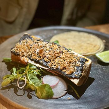 Claro - Tuetano de Mole Negro - Grilled fish with a crunchy topping is served on a black plate, accompanied by sliced radishes, greens, and a lime wedge. - Mexican Near Me - Brooklyn, New York
