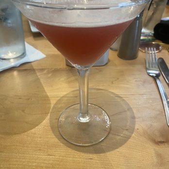 Jackson Square Grill - Pomegranate martini - A pinkish cocktail is served in a clear, stemmed martini glass on a wooden table. The drink appears slightly frothy on top. The background includes water glasses, a napkin, utensils, and condiments, indicating a dining setting. - Seafood Near Me - Columbus, Mississippi