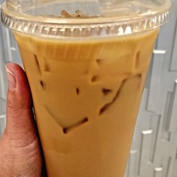 Cold Brew - A person holds a large transparent plastic cup containing iced coffee with milk and ice cubes. The cup has a clear plastic lid, and the person's hand is visible, gripping the cup. The background features gray lockers with a textured surface. The image focuses on the beverage and the hand holding it, with no additional objects or people present.