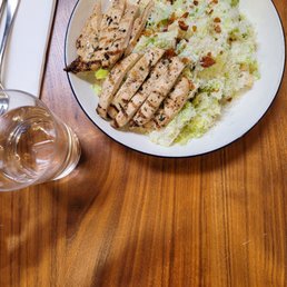 Caesar Salad - A plate of Caesar salad features sliced grilled chicken breast, chopped romaine lettuce, croutons, and a halved boiled egg with a yellow yolk. The salad is presented on a white dish with a black rim, placed on a wooden table. Nearby, there is a glass of water and a set of utensils on a napkin. The setting suggests a casual dining environment.