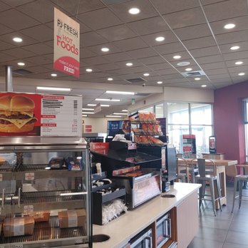 Pilot Travel Center - Other sandwiches , corn dogs condiments. - The image shows the interior of a fast-food restaurant with a ceiling fitted with multiple recessed lights and air vents. A counter displays food items and a menu board listing breakfast, burgers, hot snacks, and combo deals. Behind the counter, there are shelves with packaged food. To the right, there is a seating area with tables and chairs near large windows that let in natural light. The overall setting appears clean and organized, with a modern design. - Gas Stations Near Me - Rochester, Indiana