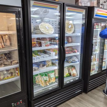 By Brazil - Lots of Brazilian staples, from toiletries to food, including frozen pizza, frozen salgadinhos, and chilled requeijão - Multiple glass-door refrigerators in a grocery store contain frozen foods, snacks, and beverages. The shelves hold items like frozen pizza, salgadinhos, and drinks. A customer wearing a blue jacket is seen browsing the products. The store has a clean, organized appearance with a wooden floor and colorful signage on top of the refrigerators. - Imported Food Near Me - Silver Spring, Maryland