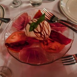 A glass plate holds slices of prosciutto, tomato, and mozzarella cheese, arranged around a ball of mozzarella topped with a basil leaf and drizzled with balsamic glaze. The plate is on a white tablecloth with silverware, wine glasses, and a water glass visible in the background, indicating a formal dining setting.