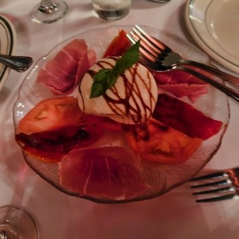 Bamonte's - A glass plate holds slices of prosciutto, tomato, and mozzarella cheese, arranged around a ball of mozzarella topped with a basil leaf and drizzled with balsamic glaze. The plate is on a white tablecloth with silverware, wine glasses, and a water glass visible in the background, indicating a formal dining setting. - Italian Near Me - Brooklyn, New York