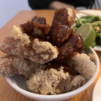 Chiko - Fried chicken! - The image shows a small white bowl filled with fried chicken pieces that have a crispy, breaded coating. The chicken is topped with a dark, glossy sauce. In the background, there is a lime wedge and some green vegetables, possibly herbs or salad. The setting appears to be a restaurant table with a wooden surface, and a person wearing a black shirt is partially visible behind the food. - Szechuan Near Me - Brooklyn, New York