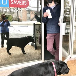 A person wearing a black face mask, glasses, a maroon beanie, a navy blue zip-up jacket, and maroon pants stands at the entrance of a pharmacy, holding a piece of paper and keys. Two black dogs are outside the door, one near the person and the other closer to the camera. A woman in a blue jacket is taking a photo from outside the store, and a woman in glasses and a mask is visible through the glass door. The background shows a street with some snow and trees.