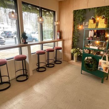 LiBre Dessert Cafe - Window seating and merchandise area. - The image shows a section of a café interior with large windows allowing natural light, a high counter with pink cushioned stools, pendant lights hanging from the ceiling, and a display shelf with various products. The background includes a green wall with decorative lighting and a floral arrangement, creating a modern and inviting atmosphere. - Coffee & Tea Near Me - Hamburg, New Jersey