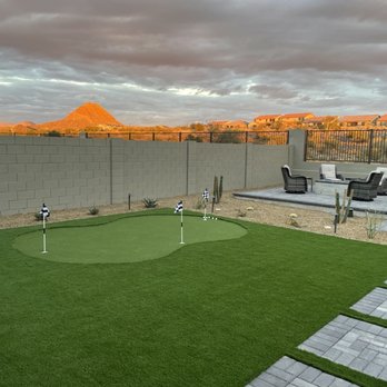 Diamond Cut Landscaping - a backyard putting green - The image shows a backyard with a synthetic grass putting green, marked by small flags, adjacent to a paved patio area with outdoor seating including chairs and a table. The yard is enclosed by a concrete block wall, with desert plants and cacti along the perimeter. In the background, there are houses and a mountain range under a cloudy sky, suggesting a desert or southwestern location. - Landscaping Near Me - Phoenix, Arizona