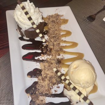 Square Cafe - Hanover - Chocolate Lave Cake with Vanilla Ice Cream and Heath Bar Crumble - A dessert plate features a slice of chocolate cake topped with whipped cream and a decorative chocolate stick. The plate also includes a scoop of vanilla ice cream, drizzled caramel and chocolate sauces, and crushed nuts or cookie crumbs. The presentation is on a white rectangular dish, placed on a restaurant table with a textured placemat and cutlery nearby. - New American Near Me - Hanover, Massachusetts