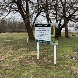 A sign in a grassy area near leafless trees welcomes visitors to the World's Largest Golf Tee in Casey, IL, with parking and business details.