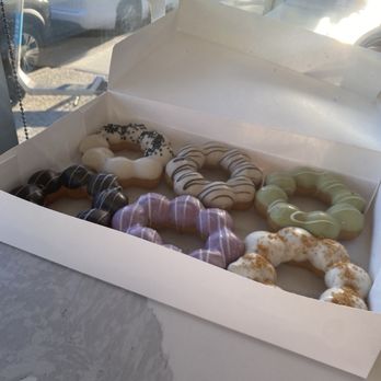 Croffle House - Mochi donuts - A white rectangular box holds several mochi donuts with various flavors and toppings, including chocolate, vanilla, and matcha, some with sprinkles or dusting. The box is on a table next to a window, with sunlight illuminating the scene and a view of parked cars outside. A vase with sunflowers is visible in the background. - Bakeries Near Me - Flushing, New York