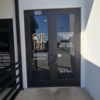 Ovr - Eze - Entrance - The image depicts the entrance of Ovr Eze, a restaurant located in Amarillo, TX. The storefront features glass double doors with business signage, including the restaurant's name and operating hours. The doors are framed in dark material, and the surrounding wall is painted white. A small portion of the exterior sidewalk and a railing are visible on the left side of the image. - Breakfast & Brunch Near Me - Amarillo, Texas