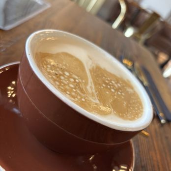 Kafeina Kafe - A close-up of a coffee cup with latte art on a wooden table in a cafe setting, with chairs and a laptop in the background. - Coffee & Tea Near Me - Spring, Texas