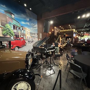 Jack’s Garage - The image shows the interior of a restaurant or bar decorated with vintage cars, including a black and a brown car, and a grand piano. A mural depicting a street scene with buildings and cars is visible on the wall. The space has dim lighting, with some string lights and spotlights, and several people are seated or standing, engaging in activities. The setting appears lively and themed with a nostalgic ambiance. - Museums Near Me - Temecula, California