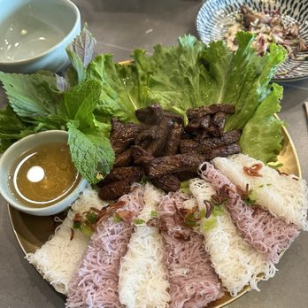 Bacogai - Beef Luc Lac - A plate displays Vietnamese rice noodle rolls, some with a pinkish hue, garnished with fried shallots and herbs. In the center, there are grilled beef pieces on a bed of lettuce and herbs, accompanied by a small bowl of dipping sauce. The presentation is on a round, gold-colored tray, with additional dishes and utensils visible in the background. - Hot Pot Near Me - Alameda, California
