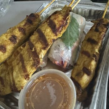 Coconut Thai Cafe - Chicken A1. Grilled Satay Chicken, A4. Veggies Rolls - Three grilled satay chicken skewers with grill marks, served with a dipping sauce and a spring roll wrapped in plastic with a leaf garnish. - Thai Near Me - Wellesley, Massachusetts