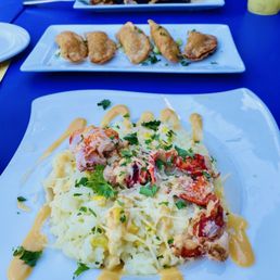 Lobster risotto, Buffalo empanadas, and marinated flank steak. - A plate of lobster mac and cheese topped with chopped parsley is in the foreground, featuring creamy cheese sauce and lobster pieces. In the background, two rectangular plates hold fried appetizers, possibly empanadas or spring rolls, garnished with herbs. The setting appears to be a casual dining restaurant with a blue table surface, and a drink with ice and lemon is partially visible on the right side.