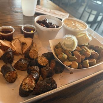 Duke City BBQ - 3 meat combo - A metal tray contains sliced smoked chicken, grilled sausage, fried okra, and small containers of barbecue sauces. There are also sides of baked beans and coleslaw with pineapple. The tray is placed on a wooden table in a restaurant with a modern interior and large windows. - Barbeque Near Me - Albuquerque, New Mexico