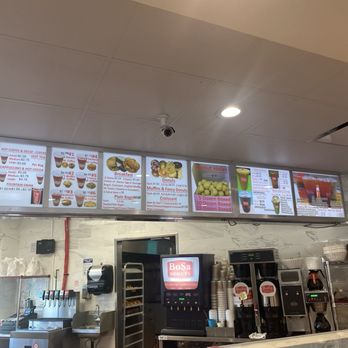 BoSa Donuts - The image shows the interior of BoSa Donuts, including a digital menu board listing donuts, breakfast items, and beverages. The counter area has coffee machines, cups, and other equipment. The ceiling has a security camera and lighting fixtures, with a wall-mounted speaker and a small window or opening behind the counter. - Donuts Near Me - Tucson, Arizona