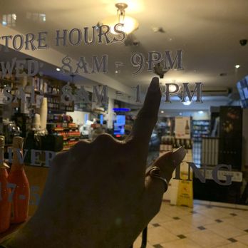 Giorgiana's - a hand pointing at the store hours window - A person’s hand, wearing a ring, is pointing at the store hours written on a glass door or window of a retail establishment. The store hours indicate opening times from 8 AM to 9 PM on Wednesday and 8 AM to 1 PM on Saturday. The interior of the store, visible through the glass, shows shelves stocked with various products, and the lighting inside is bright, contrasting with the darker exterior environment. - Beer, Wine & Spirits Near Me - Boston, Massachusetts