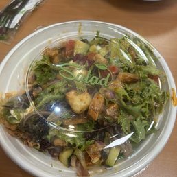 Bbq Arcadian mix salad - A mixed salad with various greens, chopped vegetables, and toppings is contained in a clear plastic container with a lid labeled 'My Salad.' The container is placed on a wooden table, with another bowl of salad and some utensils visible in the background.