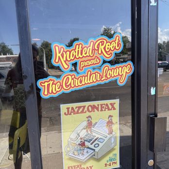 Circular Lounge - Cool logo! - The glass door displays two stickers: one with the text 'Knotted Root presents The Circular Lounge' in colorful, stylized lettering, and another poster advertising 'JAZZ ON FAX' every Sunday from 2-4 PM, featuring an illustration of a vintage telephone with musicians. The reflection of the street and a person taking the photo are visible in the glass. - Lounges Near Me - Denver, Colorado