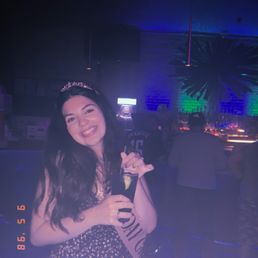 A woman with long dark hair, wearing a tiara and sash, smiles while holding a drink in a nightclub with colorful lighting.