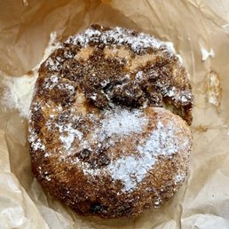 Stuff French Toast - A fried doughnut with a dark, crispy surface is shown, dusted with powdered sugar. The doughnut appears to have a rough texture with some darker spots, indicating a fried preparation. It is placed on crumpled brown parchment paper, which is used as a wrapper or serving surface.