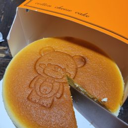 Original cheesecake - A round cheesecake with a bear design engraved on top, partially sliced, in an orange box labeled 'Mr. Bear' and 'cotton cheese cake.'