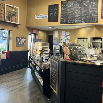 Coronation Cafe - The image shows the interior of Coronation Cafe, featuring a black counter with a glass display case and a staff member preparing orders. Behind the counter, there are menu boards listing breakfast, lunch, and beverage options. A sign reading 'Order Here' is mounted on the wall. The cafe has large windows allowing natural light, a seating area with cushions, and decorative wall accents. The overall setting is cozy and inviting, with a warm color palette and wooden flooring. - Coffee & Tea Near Me - Amherst, Massachusetts