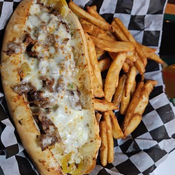 Harmon's Pub - Philly Cheesesteak - A Philly cheesesteak sandwich with melted cheese, beef, and toppings, accompanied by a portion of French fries, served on checkered paper in a casual dining setting. - Sports Bars Near Me - Canton, Ohio