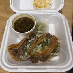Birria tacos - The image shows a foam takeout container with three birria tacos filled with shredded meat, topped with chopped onions and cilantro. Alongside the tacos, there is a cup of broth, a small container with a lime wedge, and a separate container of consommé. The setup suggests a traditional Mexican dish of birria served with broth and garnishes, placed on a wooden surface.