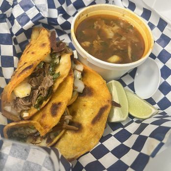 Tacos Rodriguez - Yummmmm! Delicioso - Two tacos filled with shredded beef, chopped onions, and green herbs are placed on checkered paper. A cup of broth or soup is positioned nearby, along with two lime wedges. The meal appears to be served in a casual setting, possibly at a food stand or taco truck. - Food Trucks Near Me - Phoenix, Arizona