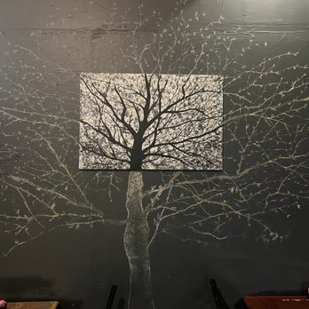 Yori - Decor- pretty - The image shows a dark-colored wall with a mural of a leafless tree painted directly onto it. The mural features a large, detailed tree with numerous branches extending outward. In the center of the mural, there is a rectangular piece of art that depicts the same tree, creating a layered visual effect. The setting appears to be indoors, with parts of furniture and electrical cords visible at the top and bottom edges of the image. - Korean Near Me - Hurst, Texas