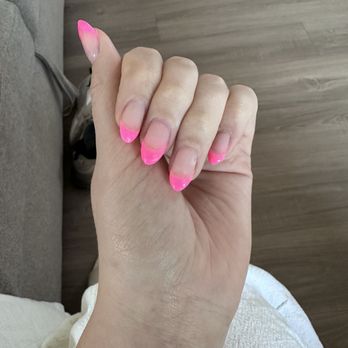 DK Nails - The image shows a hand with five fingers, each featuring a pink French manicure with bright pink tips and a natural, clear base. The hand is positioned against a background of a wooden floor and part of a sofa, with some shoes visible in the background. The nails are well-maintained and painted with a glossy finish. - Nail Salons Near Me - Austin, Texas