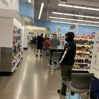Walgreens - Hour long wait - A person wearing a black face mask, black jacket with white stripes, and green pants is standing in line at a pharmacy checkout counter inside a retail store. The store has shelves stocked with various products, and other customers are visible in the background. The ceiling features exposed pipes and lighting fixtures, and there are chairs and signage indicating pharmacy services. - Drugstores Near Me - Concord, California