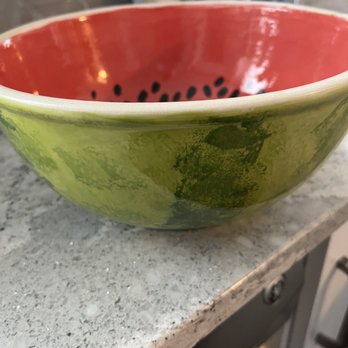 Color Me Mine - A ceramic bowl with a watermelon-themed design, showing a green exterior with darker green patches and a red interior decorated with black seed patterns. The bowl is positioned on a speckled gray kitchen countertop, with kitchen appliances and items visible in the background. - Paint-Your-Own Pottery Near Me - Glenview, Illinois