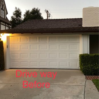 I Do Remodeling - Driveway before - The image shows a residential garage with a white sectional garage door and a concrete driveway leading up to it. The driveway has visible cracks and stains. To the right, there is a small green hedge and a brick pathway. The house has a brown shingle roof, and part of the neighboring house is visible on the left. The sky is overcast or at dusk, and a warm exterior light is on near the garage. The text 'Drive way Before' is overlaid in red on the lower part of the image. - General Contractors Near Me - Fullerton, California