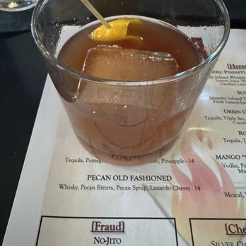 Dante's Fire - pecan old fashioned - A clear glass contains a pecan old fashioned cocktail with a large ice cube and a lemon wedge garnish, resting on a restaurant menu. The menu lists various drinks, including the pecan old fashioned, and features a section titled 'Fraud' with other cocktail options. The setting appears to be a dining or bar area. - Cocktail Bars Near Me - Tucson, Arizona