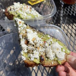 Millennial Toast - A hand holds a slice of avocado toast topped with cheese, seeds, and spices, with a plastic container of more toast in the background on a metal table.
