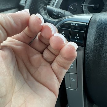 Indulgence Nails and Spa - Thick wavy nails - A close-up of a person's left hand with French manicured nails, featuring white tips and natural pink bases, is shown inside a car. The hand is near the steering wheel, which is wrapped in black leather. The dashboard and control buttons, including volume and mode controls, are visible in the background, along with part of the speedometer and other gauges. - Nail Salons Near Me - Saugus, Massachusetts