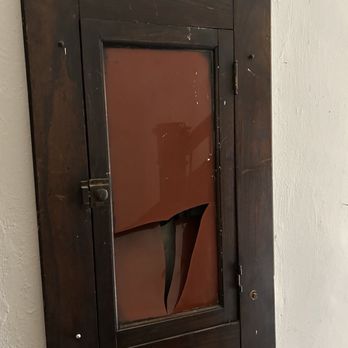 Arch Moving - broken window - A rectangular wooden cabinet with a glass front is mounted on a white wall. The glass door is broken, with a large, irregular hole and jagged edges. The cabinet appears old, with visible scratches and wear on the wood. The wall behind it is plain and textured. - Movers Near Me - Saint Ann, Missouri