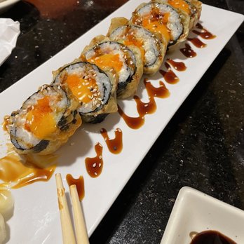 Oriental Express - Yummy Roll - A long, rectangular white plate features six sushi rolls topped with orange sauce and sprinkled with sesame seeds. The rolls are arranged in a line on a dark table, with chopsticks resting on the plate and a small dish of soy sauce nearby. The background includes a partially visible cup and a smartphone. - Chinese Near Me - Topeka, Kansas