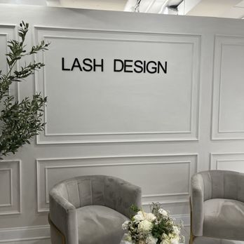Lash Design - The photo depicts the interior of Lash Design, featuring a white wall with decorative paneling and the business name in black letters. Two gray upholstered chairs with gold accents are positioned in front of the wall. A floral arrangement with white flowers and greenery is placed on a small table, along with a water bottle and other small items. The ceiling has visible white ductwork and lighting fixtures, indicating a modern, minimalist aesthetic. - Eyelash Service Near Me - Chicago, Illinois