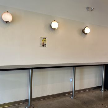 Sunright Tea Studio - A long, black counter with metal supports runs along a plain, light-colored wall in a cafe or tea shop. Three wall-mounted lights with round shades are evenly spaced above the counter. There are electrical outlets near the floor, and a small poster and a sign are visible on the wall. The setting appears clean and minimalistic, with no customers or staff present. - Bubble Tea Near Me - Fullerton, California