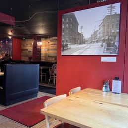 Interior - A wooden table with a red-cushioned chair is positioned near a red wall in a restaurant interior. The wall displays a large black-and-white photograph of a historic street scene with buildings and trolley tracks. The background includes additional seating, a wooden accent wall, and dim lighting, creating a warm, inviting ambiance.