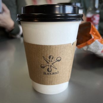 Blackjack’s - A disposable coffee cup made of paper with a black plastic lid and a brown paper sleeve displaying the BlackJacks logo, which consists of a stylized design with scissors and a needle. The cup is on a table, with a blurred background that includes a person’s arm and a snack bag, indicating a casual setting, likely inside a coffee shop or similar establishment. - Coffee & Tea Near Me - Dublin, Texas