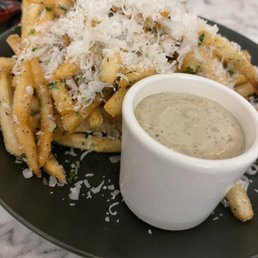 Parmesan Truffle fries - A serving of French fries is presented on a black plate, topped with grated Parmesan cheese and shredded cheese, garnished with herbs. A small white cup containing a creamy, speckled dipping sauce is placed beside the fries. The background includes a marble table surface and some out-of-focus objects, indicating a casual dining setting.